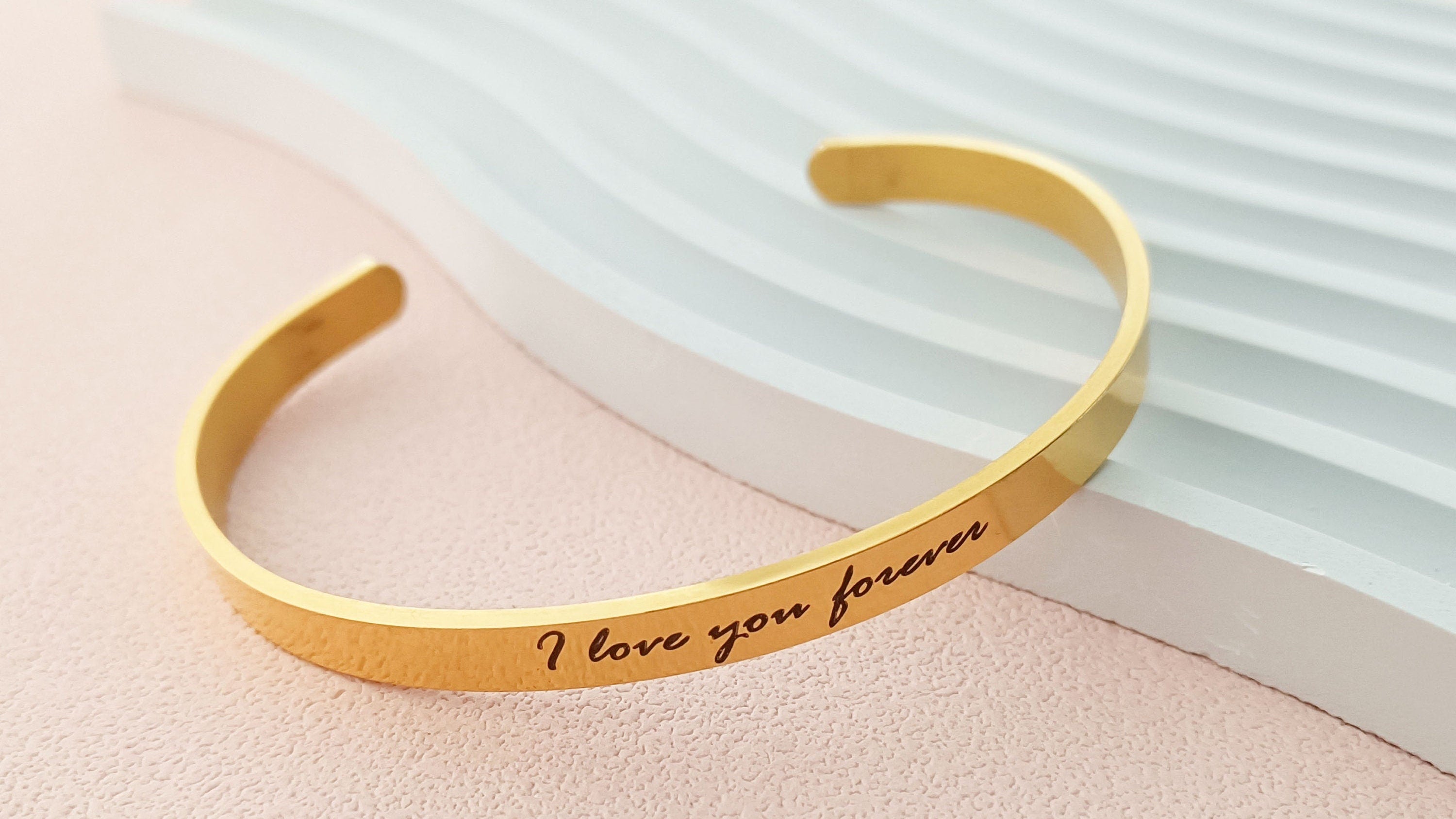 Gold bracelet with 'I love you forever' engraving on a light pink surface.