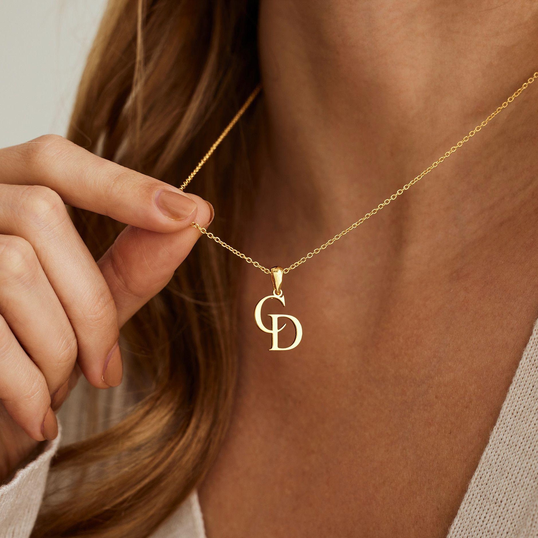 Gold necklace with a pendant featuring initials, held by a hand against a neutral background.